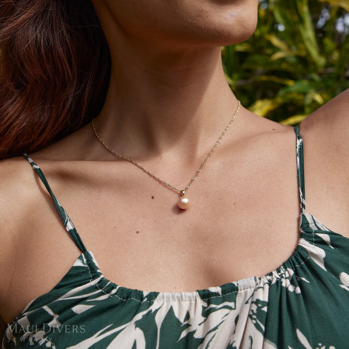 Close-up of a woman in a green top wearing a simple peach/pink pearl pendant with a 14k yellow gold bail on a singapore chain with plants in the background