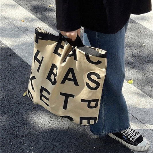 Model carring a khaki canvas tote bag with black alphabet print and double shoulder straps.