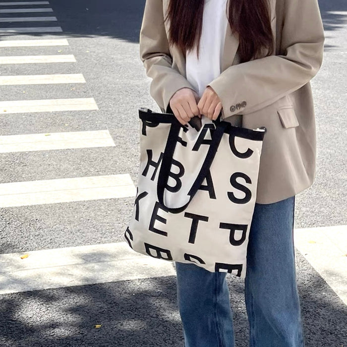 Model carring a white canvas tote bag with black alphabet print and double shoulder straps.