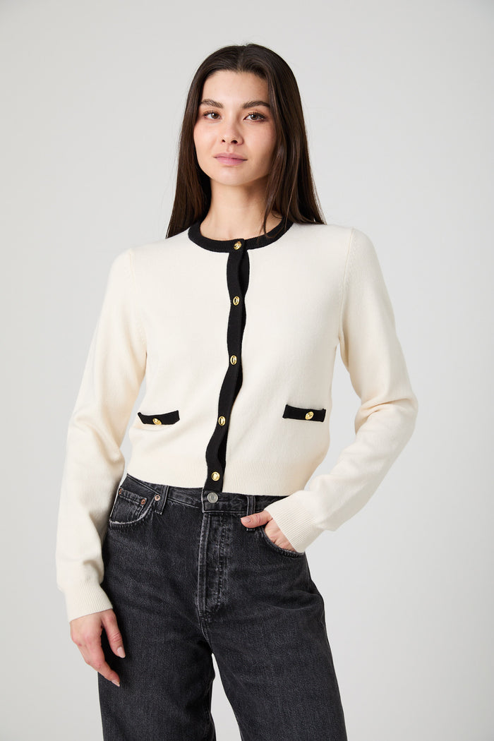 A woman modeling a cream-colored knit cardigan with black trim and gold buttons, paired with dark gray jeans, against a light gray studio background.
