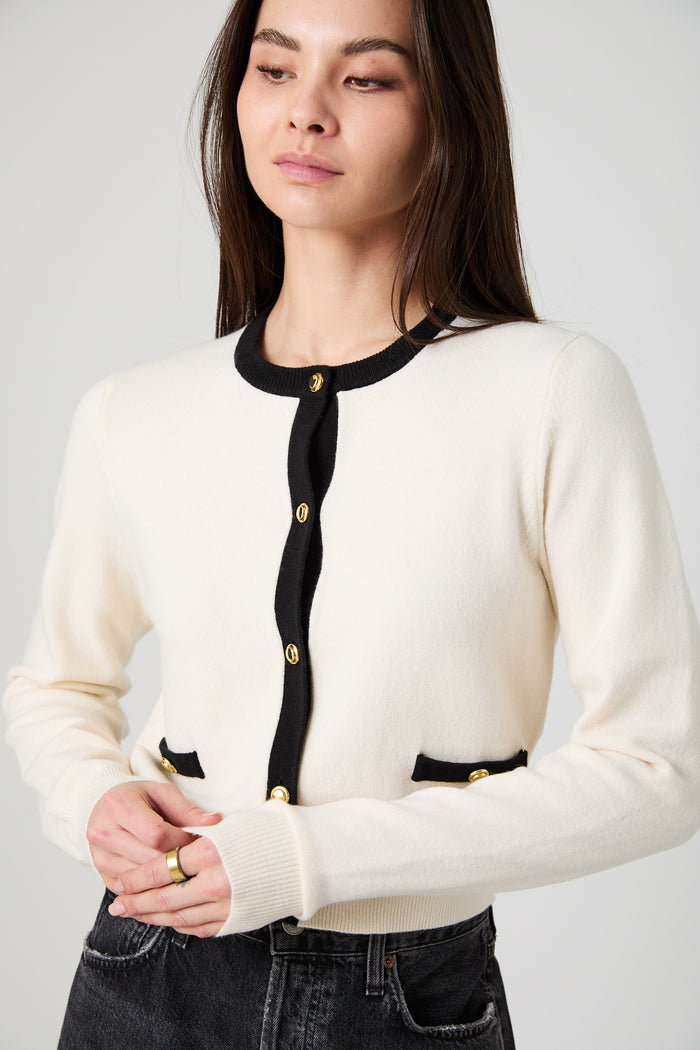 A woman wearing a cream-colored knit cardigan with black trim and gold buttons, paired with dark jeans, against a light gray studio background.