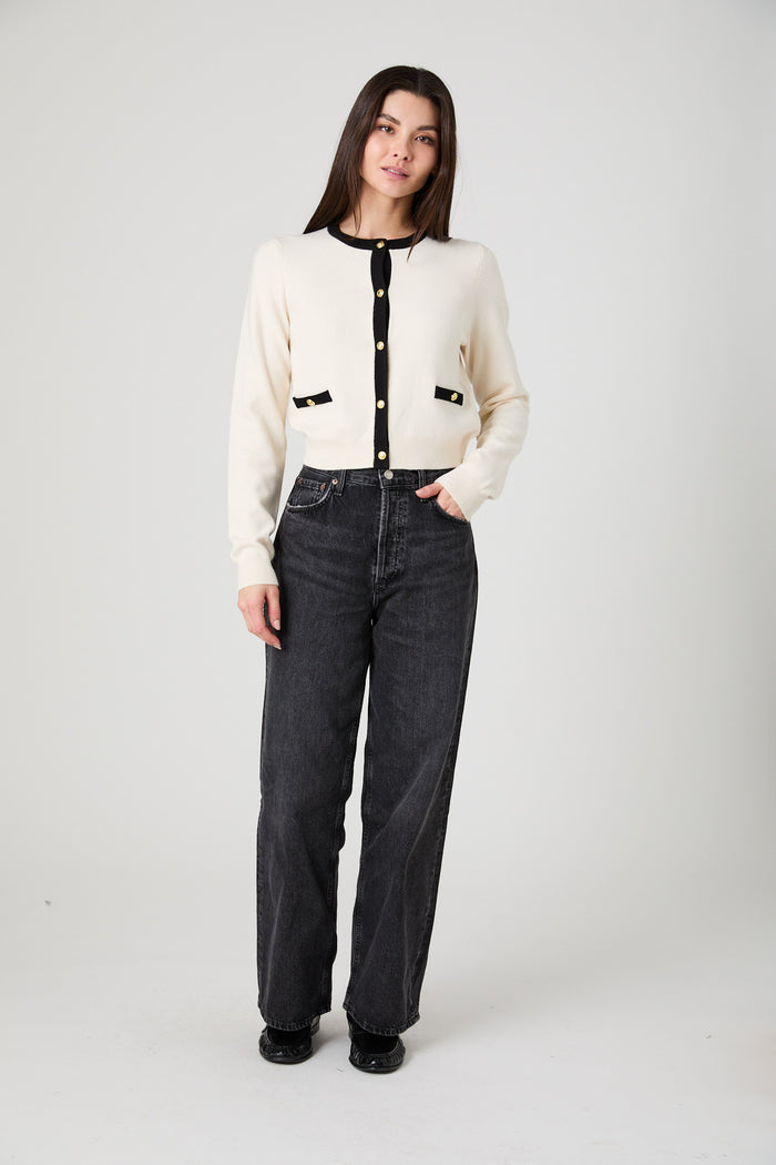 A woman modeling a cream-colored knit cardigan with black trim and gold buttons, paired with dark gray jeans, against a plain light gray studio background.