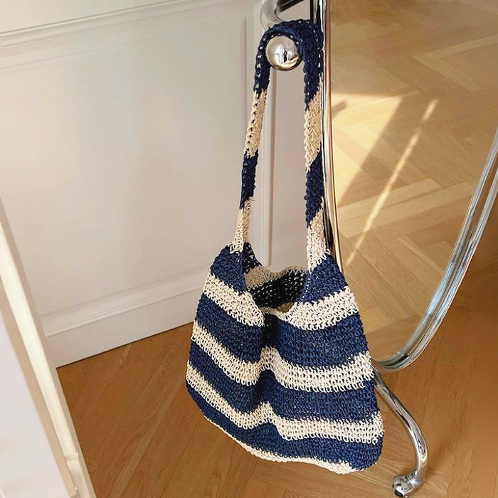 Close-up of Crochet straw tote bag with blue and natural-toned stripes, hanging from a chrome rail, ideal for errands.