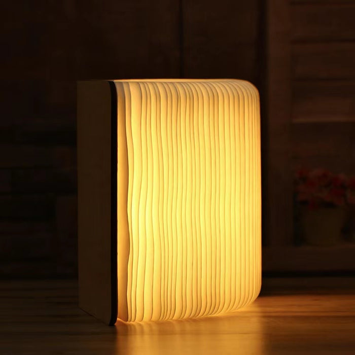 Walnut wood book lamp, open to show warm LED light, standing upright on a dark wooden surface.