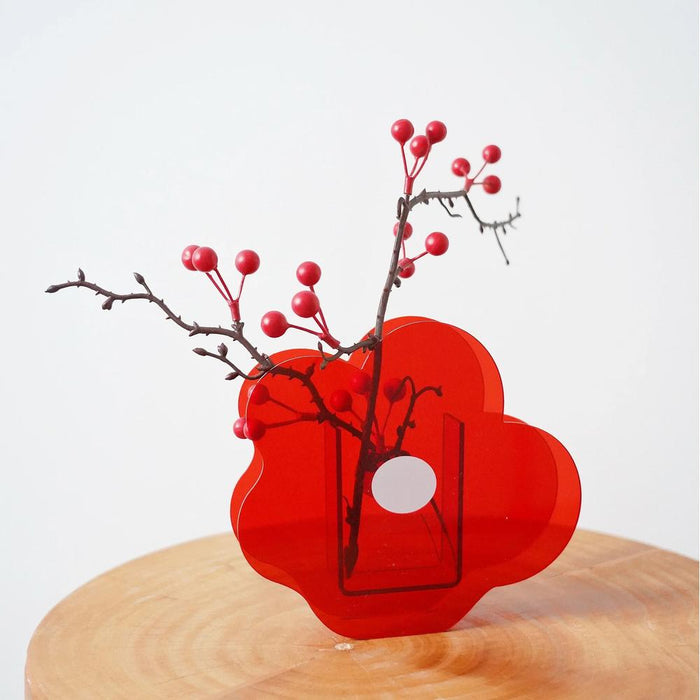 Angled view of Red acrylic flower-shaped vase with berry branch on wooden table.