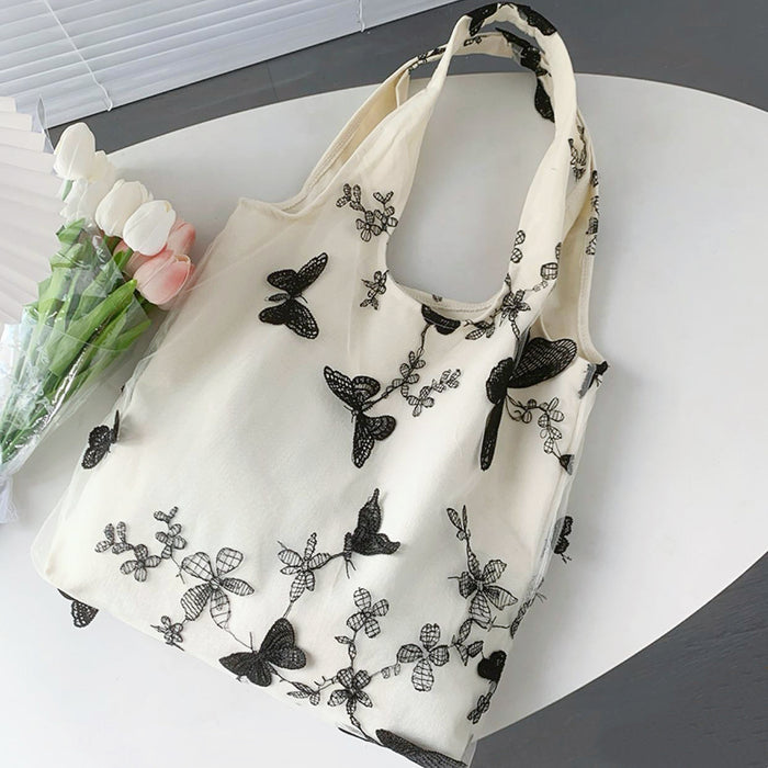 Cream canvas tote bag with black butterfly and floral embroidery, open top, shoulder strap.