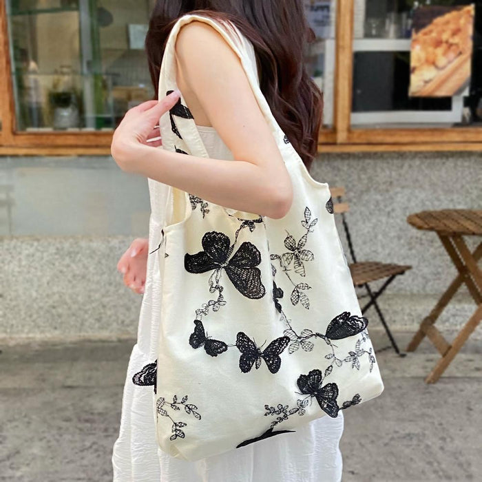 White canvas shoulder bag with black lace butterfly embroidery, held by a woman.