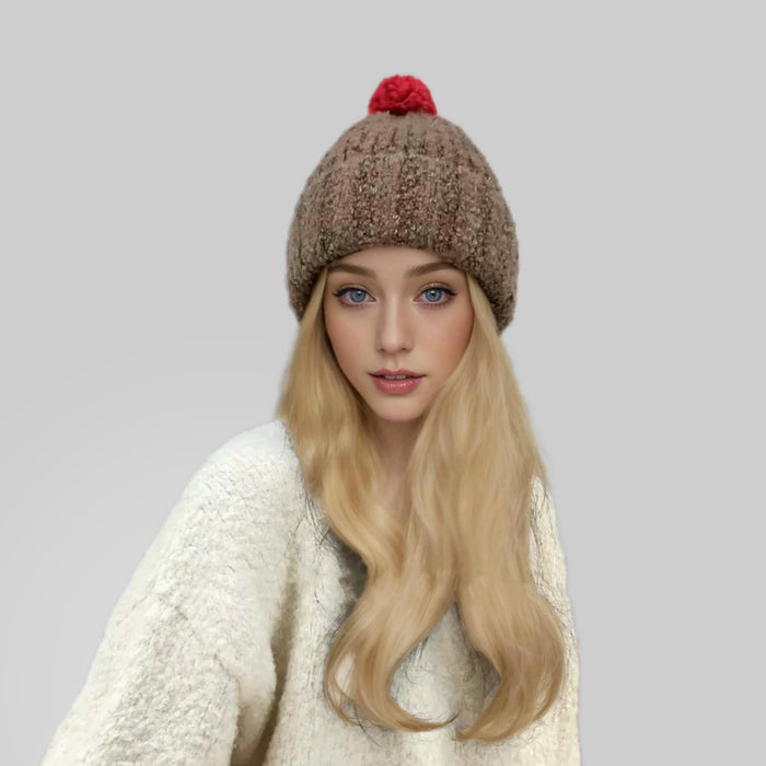 Woman wearing brown knitted pompom beanie with red accent next to a white sweater.