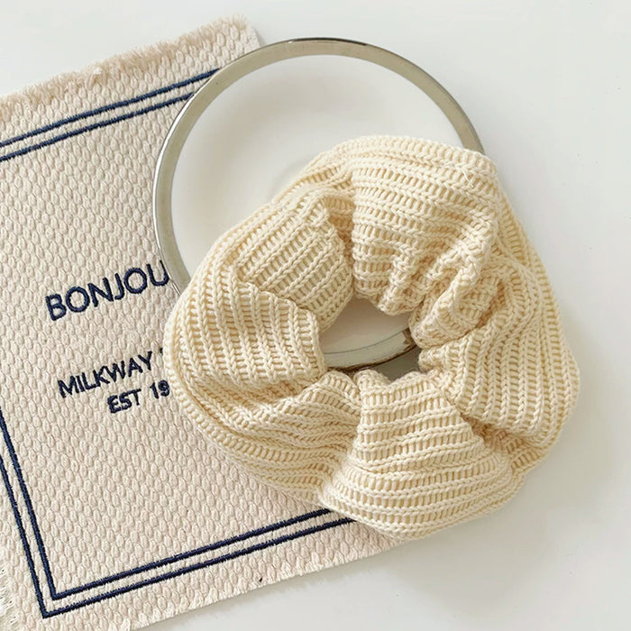 Front view of Cream knitted  scrunchie with ribbed texture, resting on a white plate and beige fabric.