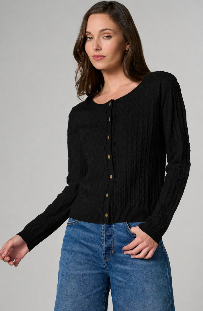 A woman wearing a black long-sleeve button-front cardigan with gold buttons and blue jeans, photographed against a plain light gray studio background.