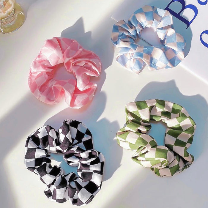 Flat lay of four satin checkerboard scrunchies in pink, blue, black, and green.