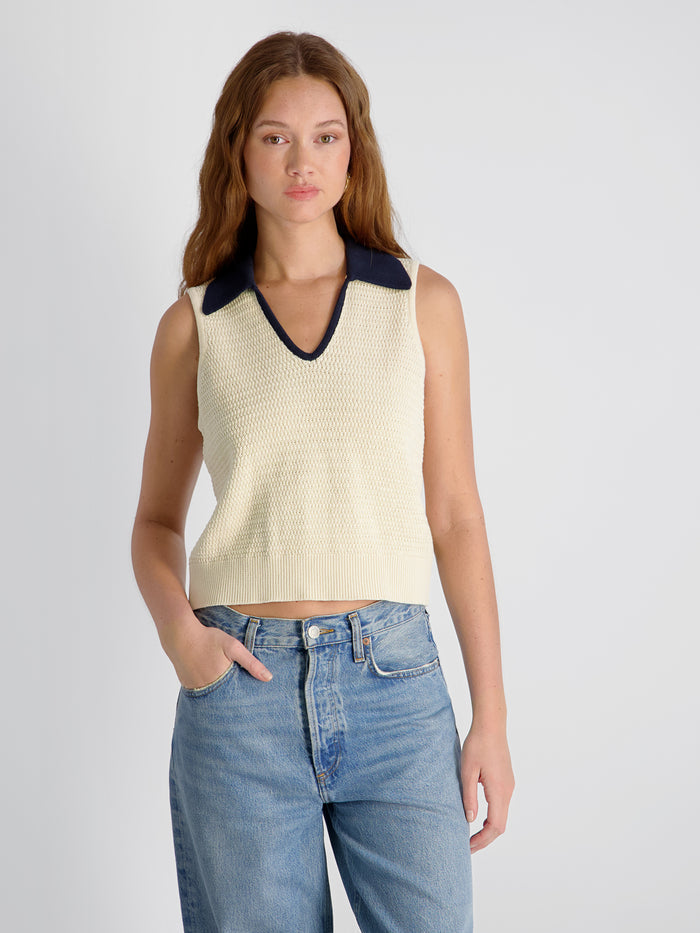Woman wearing a cream sleeveless top with a navy collar and blue jeans on a light gray background