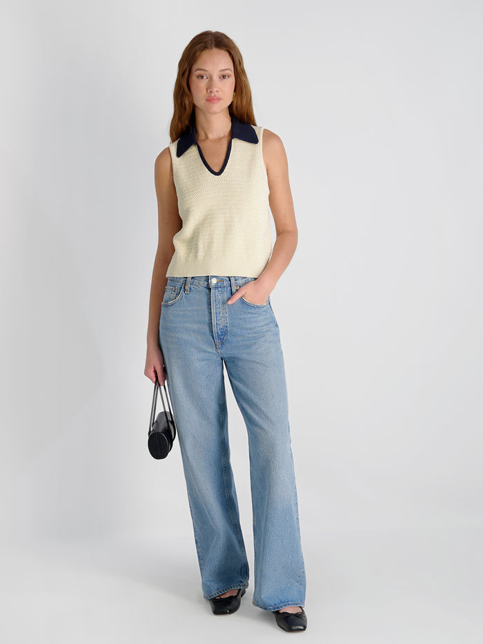 Woman wearing a sleeveless cream top with a black collar and light blue jeans on a plain background