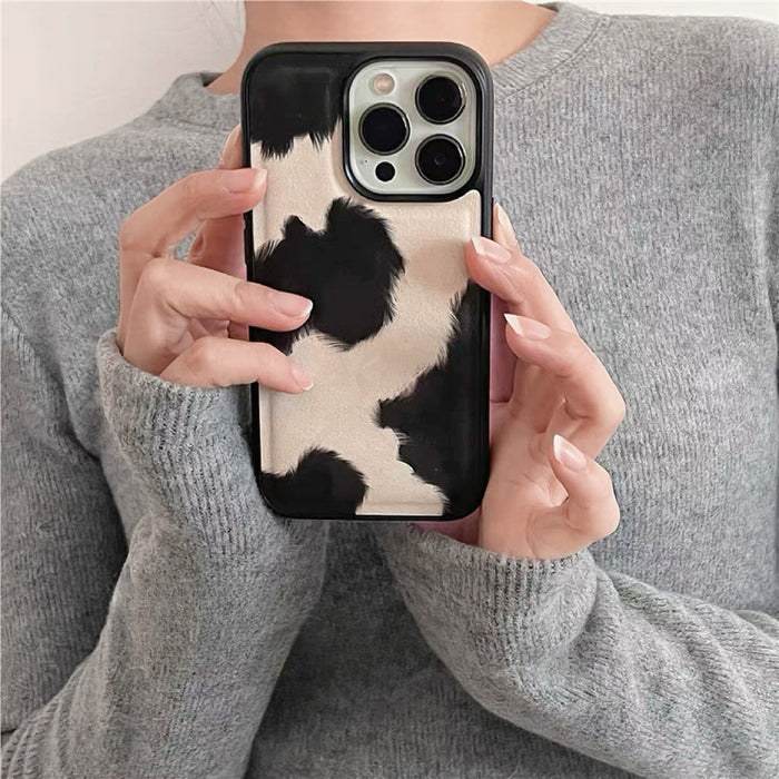 Hands in gray sweater holding black and white cow print PU leather phone case. Cozy autumn fashion vibes.