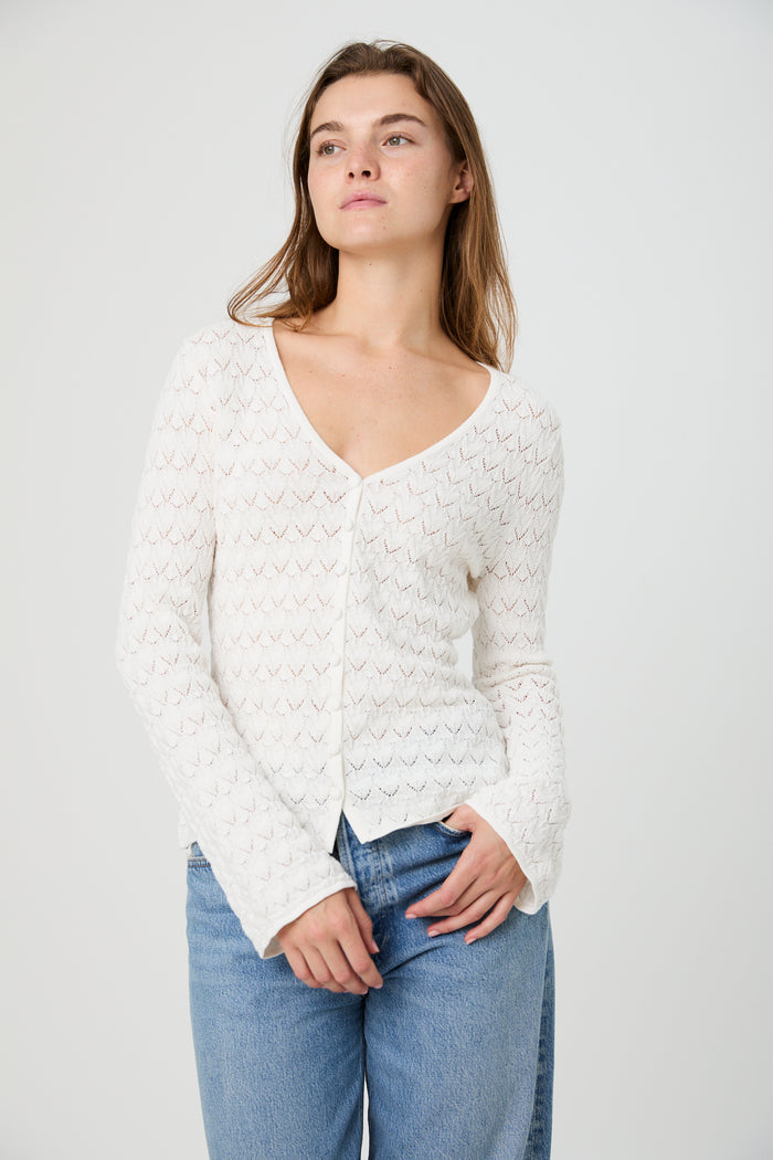 Woman wearing a white textured cardigan and blue jeans on a plain background
