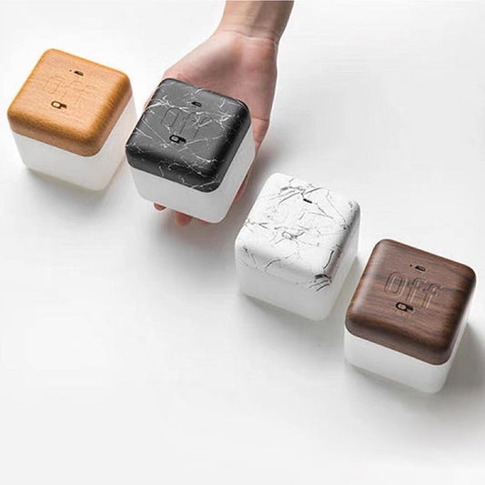 Hand holding a dark marble Flipping Cubic Timer Lamp, with beech wood, white marble, and walnut wood lamps beside it.