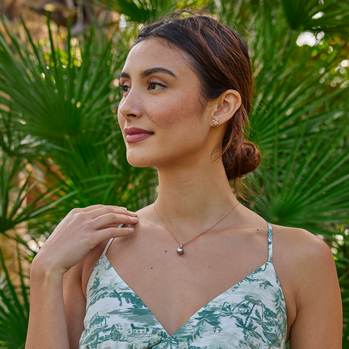 A woman wearing a lilac freshwater pearl pendant in rose gold and plumeria earrings with a blurry palm tree background.