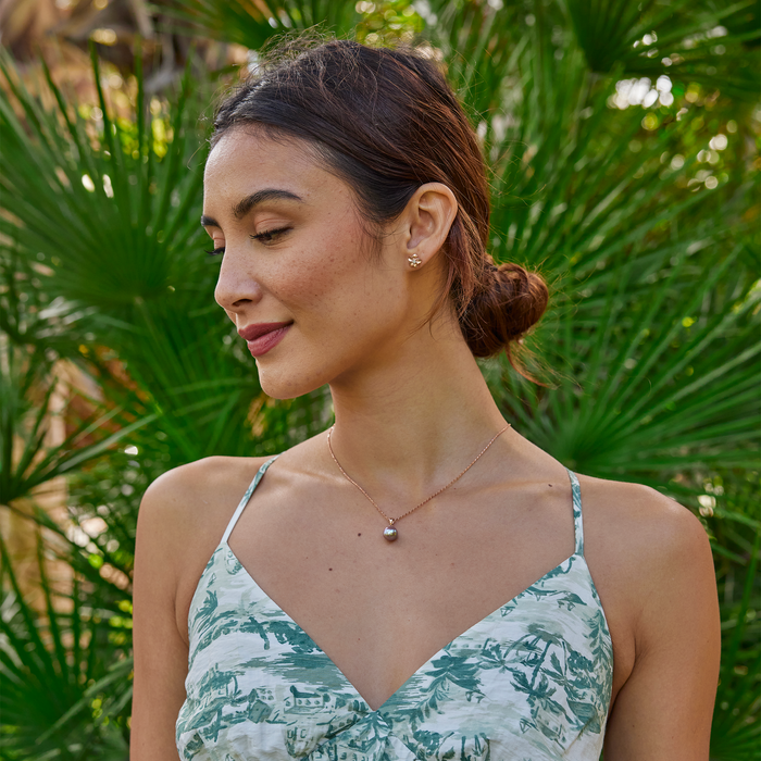A woman wearing a lilac freshwater pearl pendant in rose gold and plumeria earrings with a blurry palm tree background.