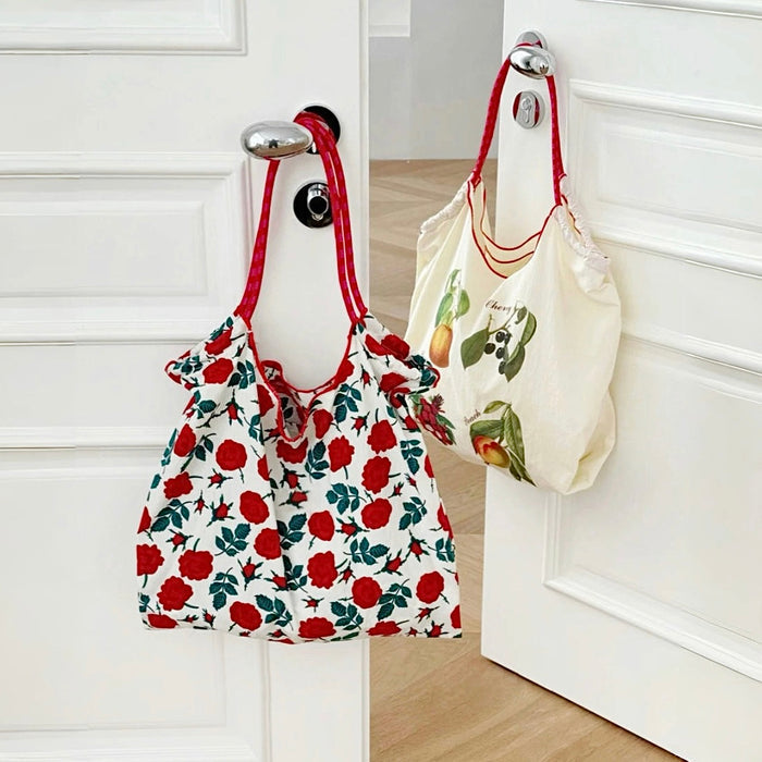 White polyester tote bag with red rose print and red straps, next to a cream fruit-print tote.