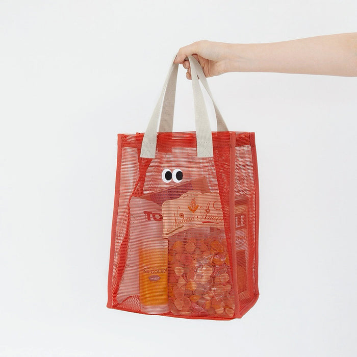Angled view of orange polyester mesh shopper bag with googly eyes, beige shoulder straps.