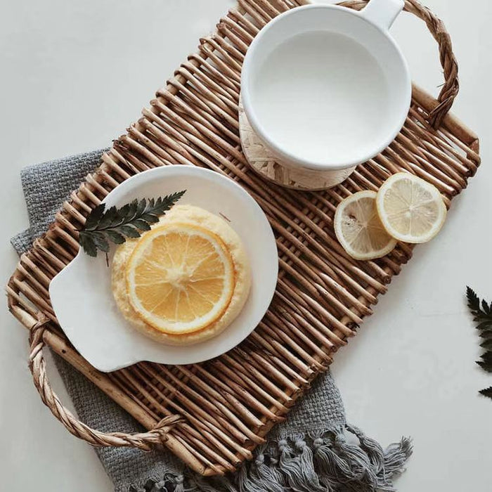 Front view of Hand-woven rattan tray with handles, holding a white mug, lemon pastry, and slices.