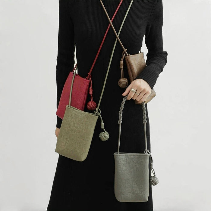 Four hanging ball leather crossbody bags in red,  brown, grey and khaki green, worn together by a person.