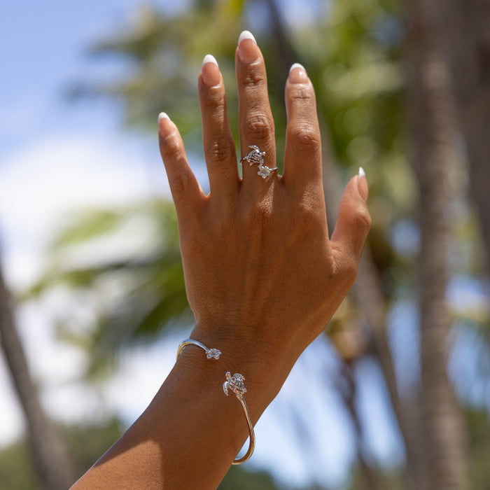 A woman's hand wearing a Honu and Plumeria Bypass Ring in Sterling Silver with blurred palms in the back
