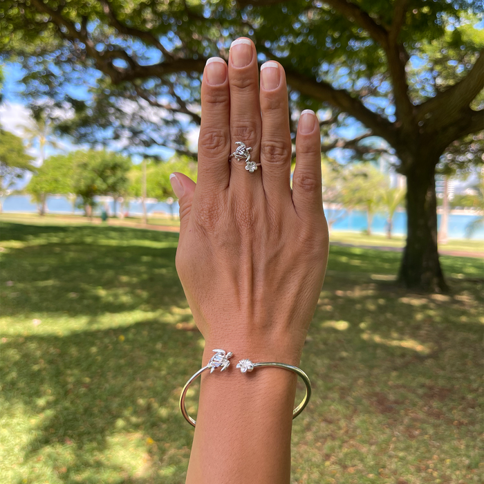A woman's hand holding a Honu and Plumeria Ring and bracelet in Sterling Silver with a beach park in the back