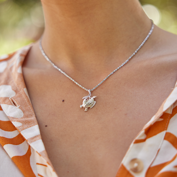 Close up of Honu Pendant / Charm in Sterling Silver on a neckline with blurred greenery in the back
