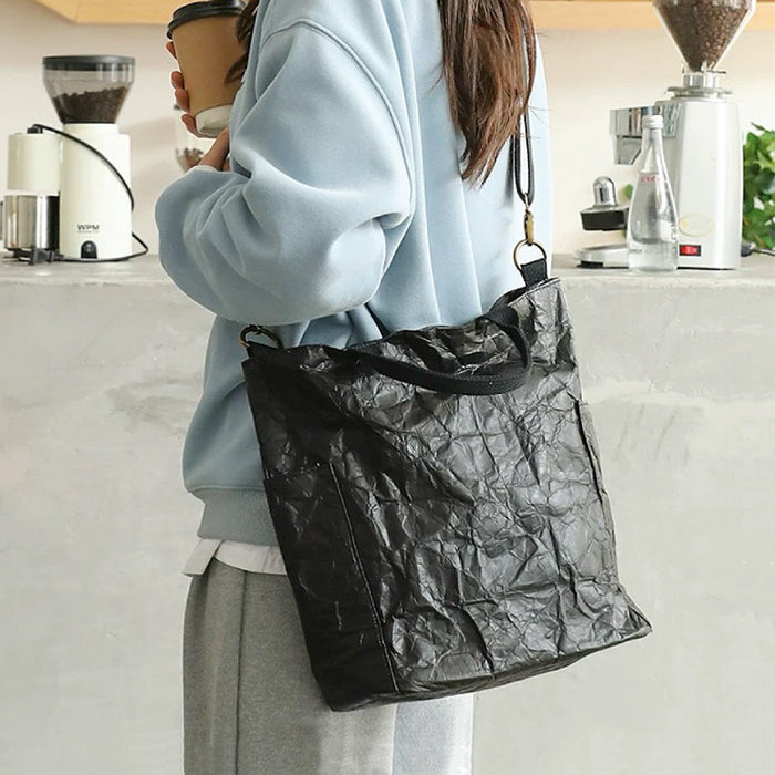 A model wearing a black kraft-paper-textured tote bag as crossbody, showcasing a modern urban explorer look.