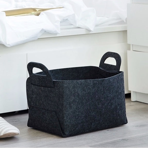 A black storage basket with handles sitting on a floor next to a white cabinet.
