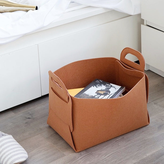 A tan felt storage basket containing books, placed next to a bed.