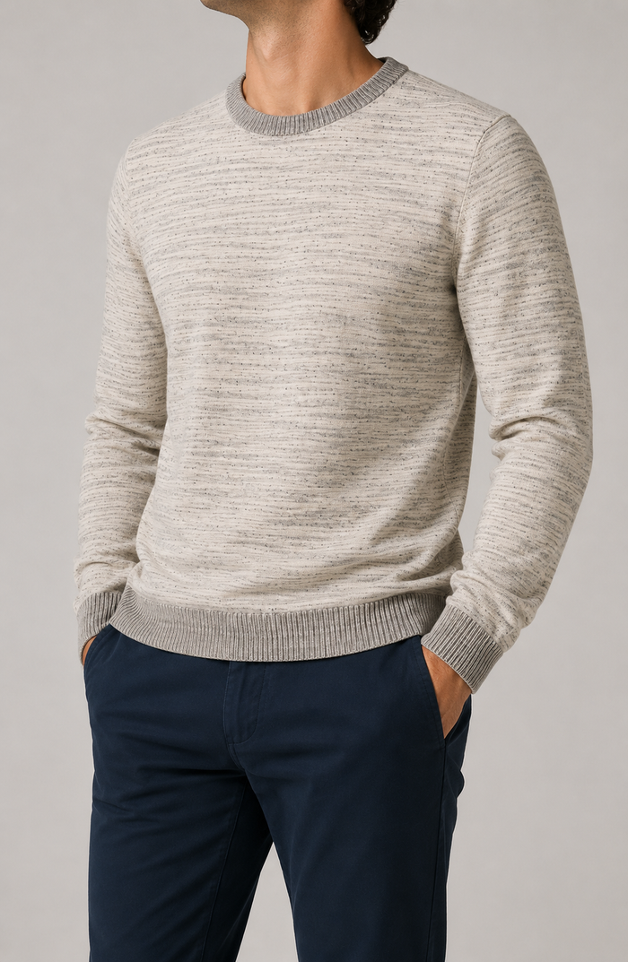 A man wearing a light beige marled knit crewneck sweater with gray ribbed trim, paired with navy blue trousers, against a neutral gray background.