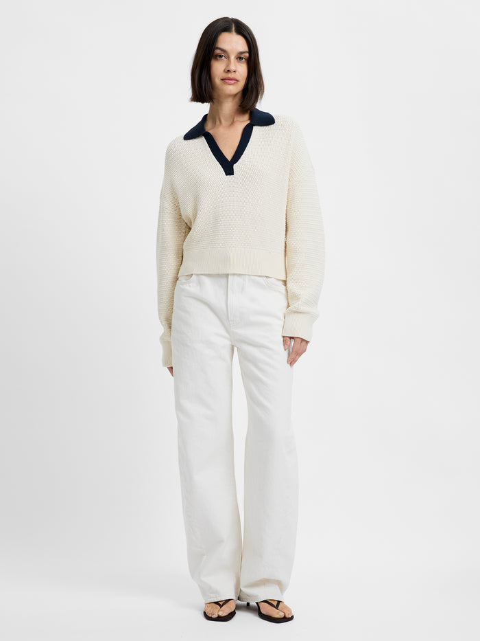 Woman wearing a cream sweater with a navy collar and white pants on a white background