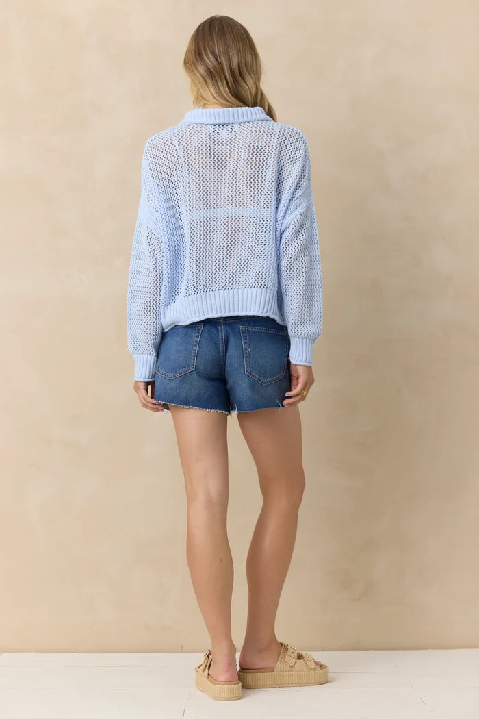 Woman wearing a light blue textured cardigan and denim shorts against a beige background