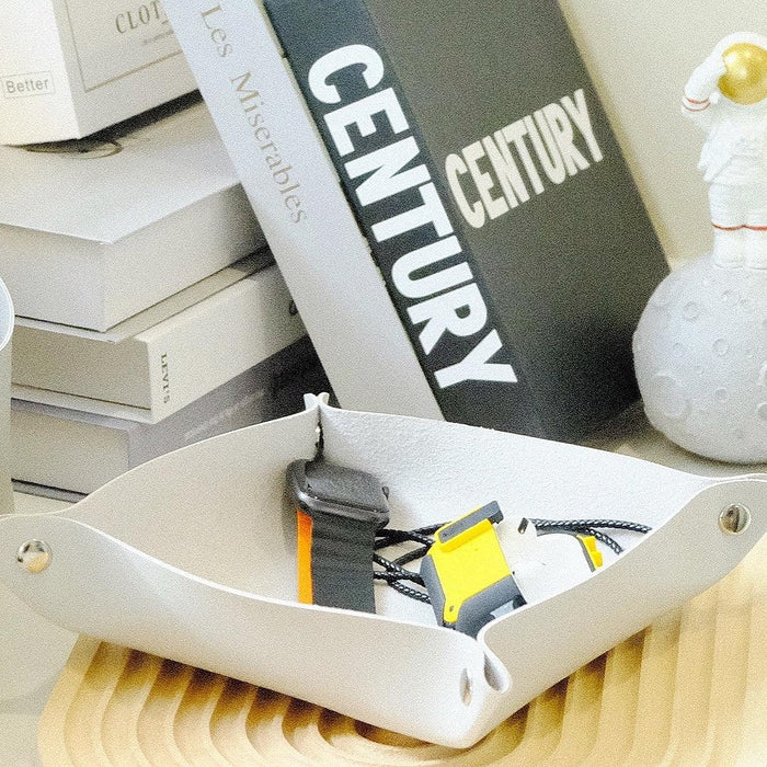 White leather tray on wooden surface holding a smartwatch and small accessories, books in background.