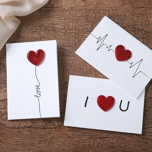 Three white paper love cards with red heart designs and black text on a wooden surface.