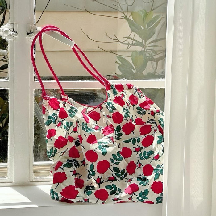 Angled view of Polyester Rose Romance Tote Bag with red rose print, red grab straps, hanging in a window.