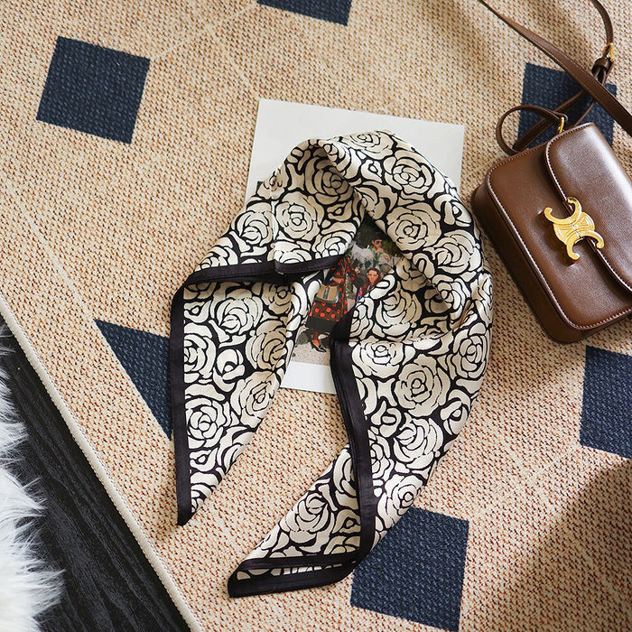 Angled view of Rose silk scarf, with black and white rose pattern, on a patterned rug, easy for travel.