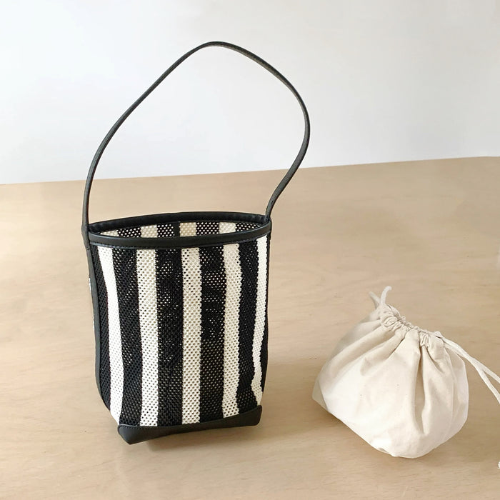 Close-up of Black and white stripe woven raffia bucket bag with PU leather trim and single strap, next to a small pouch.