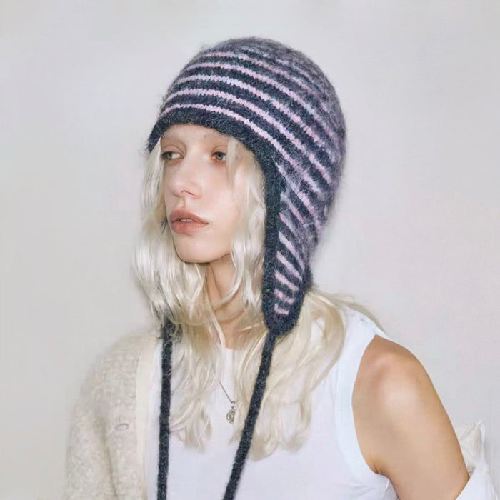 Model wearing a pink and dark grey striped knit earflap hat with braided tassel ties.