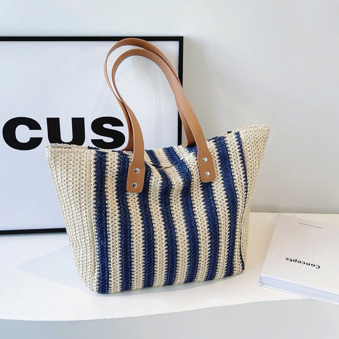 Angled view of Vertical striped straw tote bag with blue and natural stripes, brown PU leather shoulder straps.